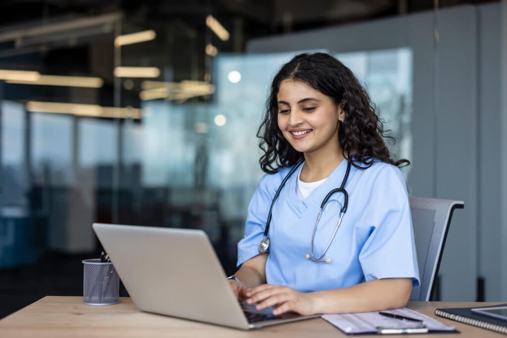 Young smiling female medical professional in scrubs and stethoscope working on a laptop at a modern clinic desk, typing and providing telehealth support and patient care remotely