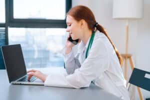 Side view of happy smiling female doctor in white coat typing on laptop keyboard and talking on mobile phone with patient during remote consultation, sitting at desk near window in sunny day.