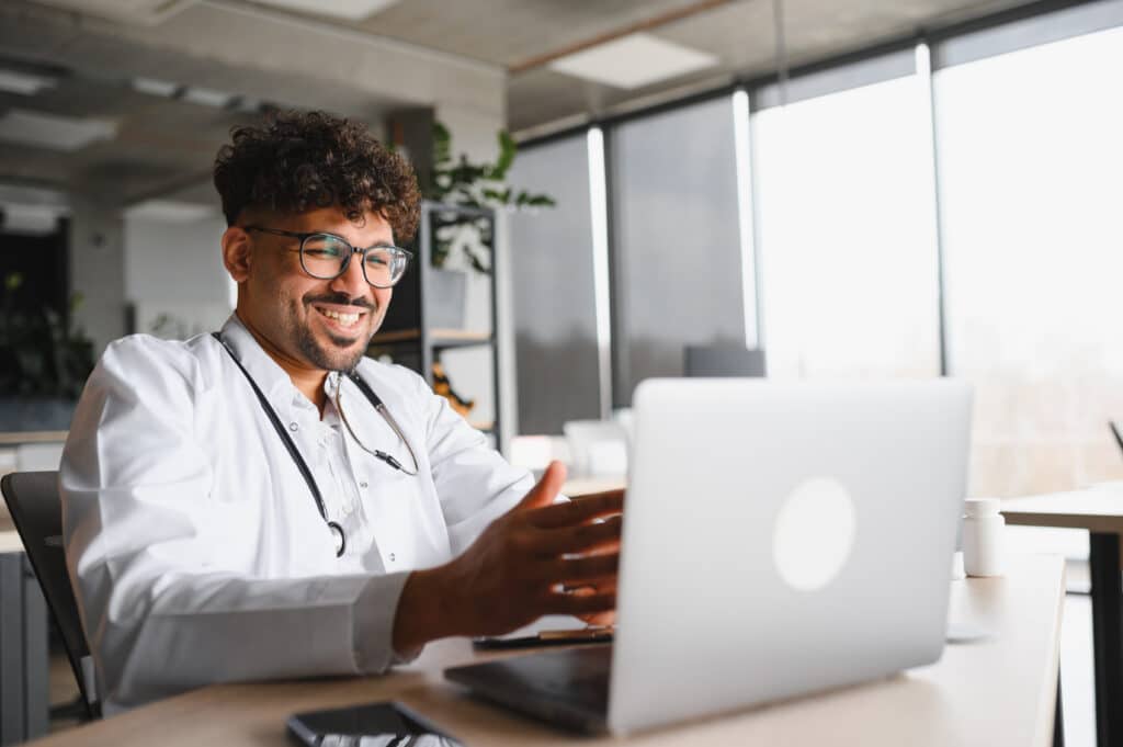 Smiling arab doctor wearing lab coat and stethoscope using laptop for video consultation in bright medical office